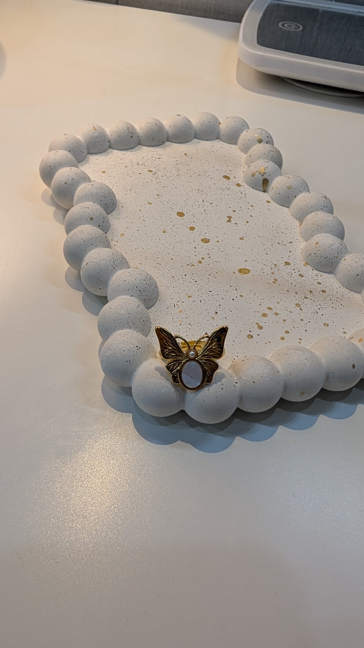 Gold-tone butterfly ring with textured white wings and a pale purple oval center, resting on a white speckled dish.