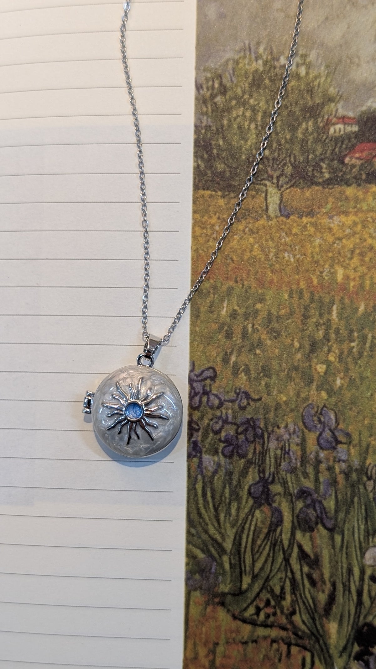 A silver-tone necklace featuring a fine chain and a round locket pendant with an engraved sunburst design and a central blue accent.