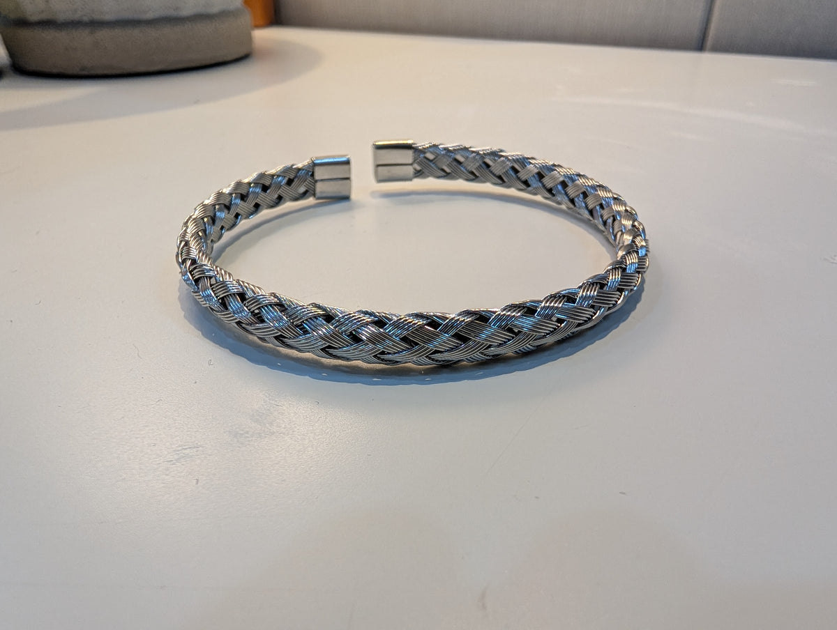 Silver-tone open cuff bracelet with a braided woven pattern and flat ends resting on a white surface.
