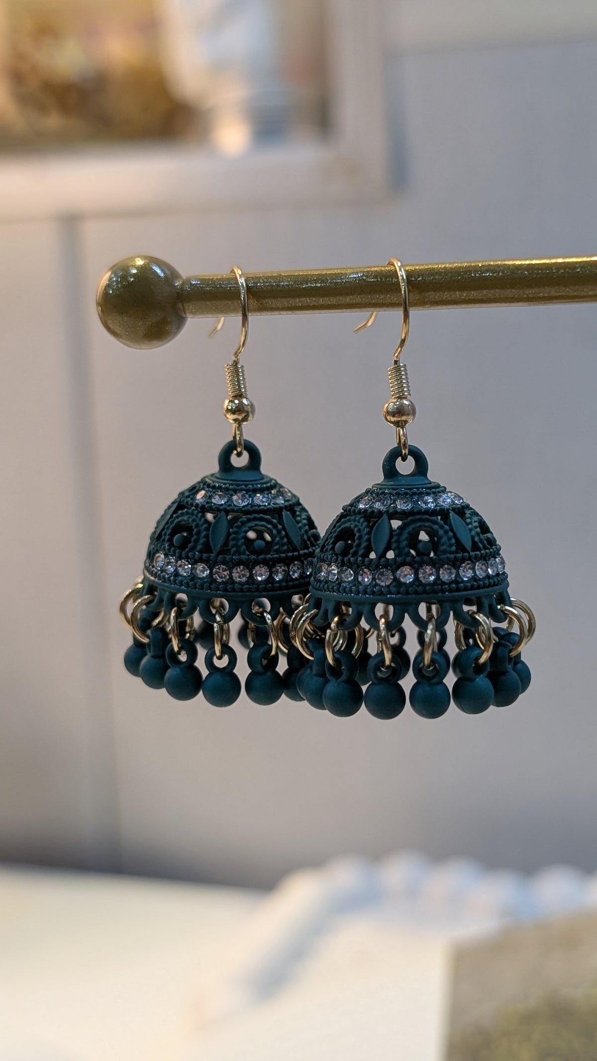 Pair of dark teal dome-shaped dangle earrings hanging from a horizontal rod, showing openwork patterns, small pale embellishments, and dangling round teal beads.