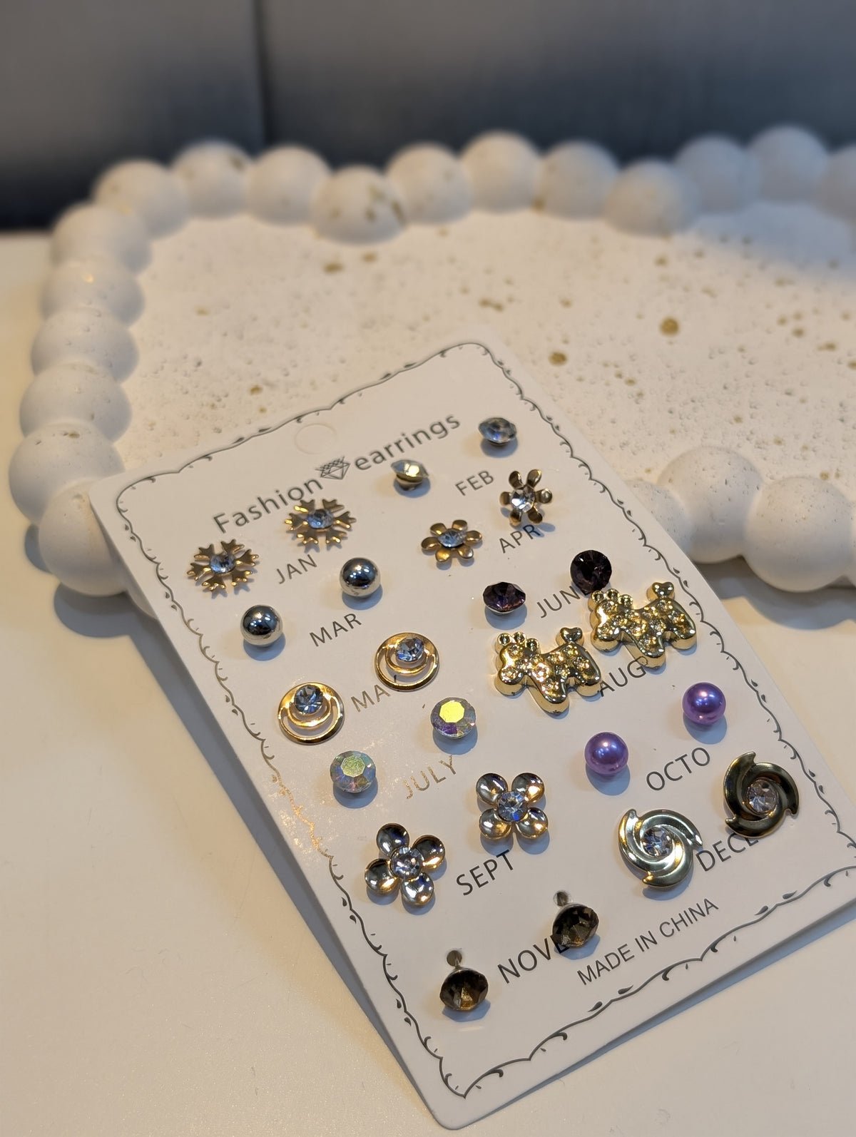 A display card holding an assortment of stud earrings with monthly labels, featuring floral, star, bow, and swirl designs with colored accents.