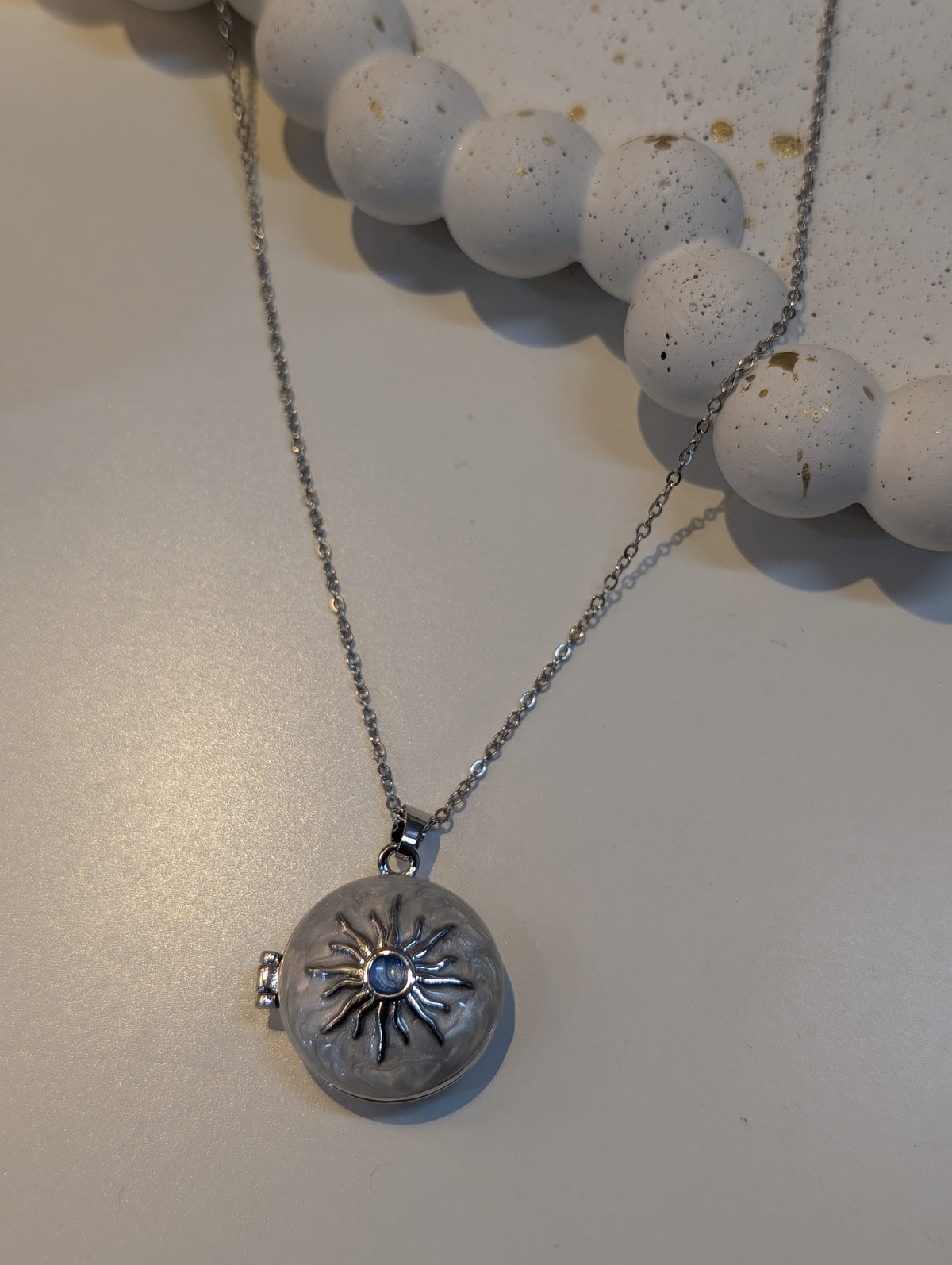 Silver-tone necklace with a fine cable chain and a round pendant decorated with a raised sunburst design around a small blue centre stone, shown on a light surface.