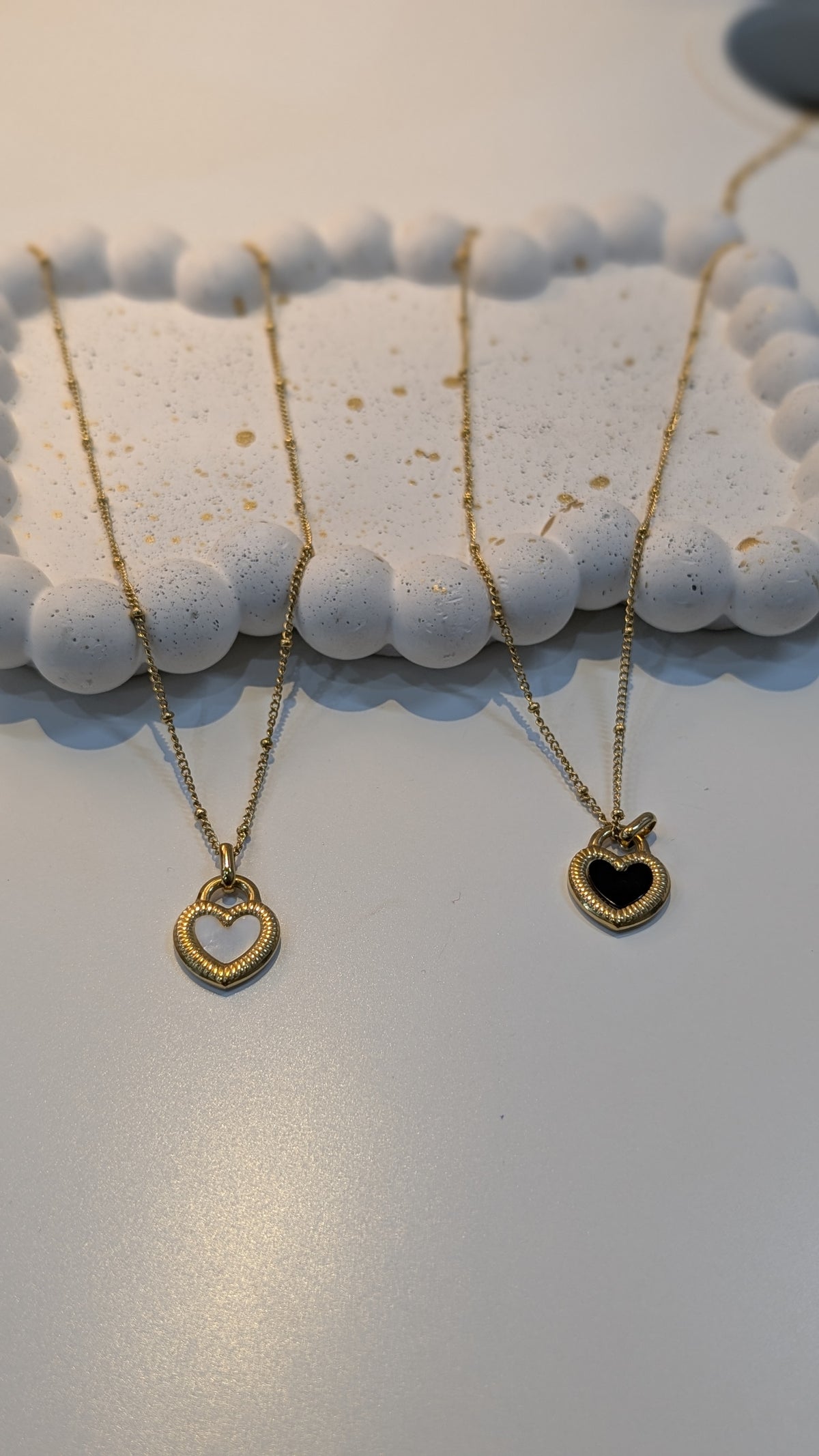 Two gold-tone box chain necklaces with heart pendants, one with a light center and one with a dark center, resting on a textured white tray.