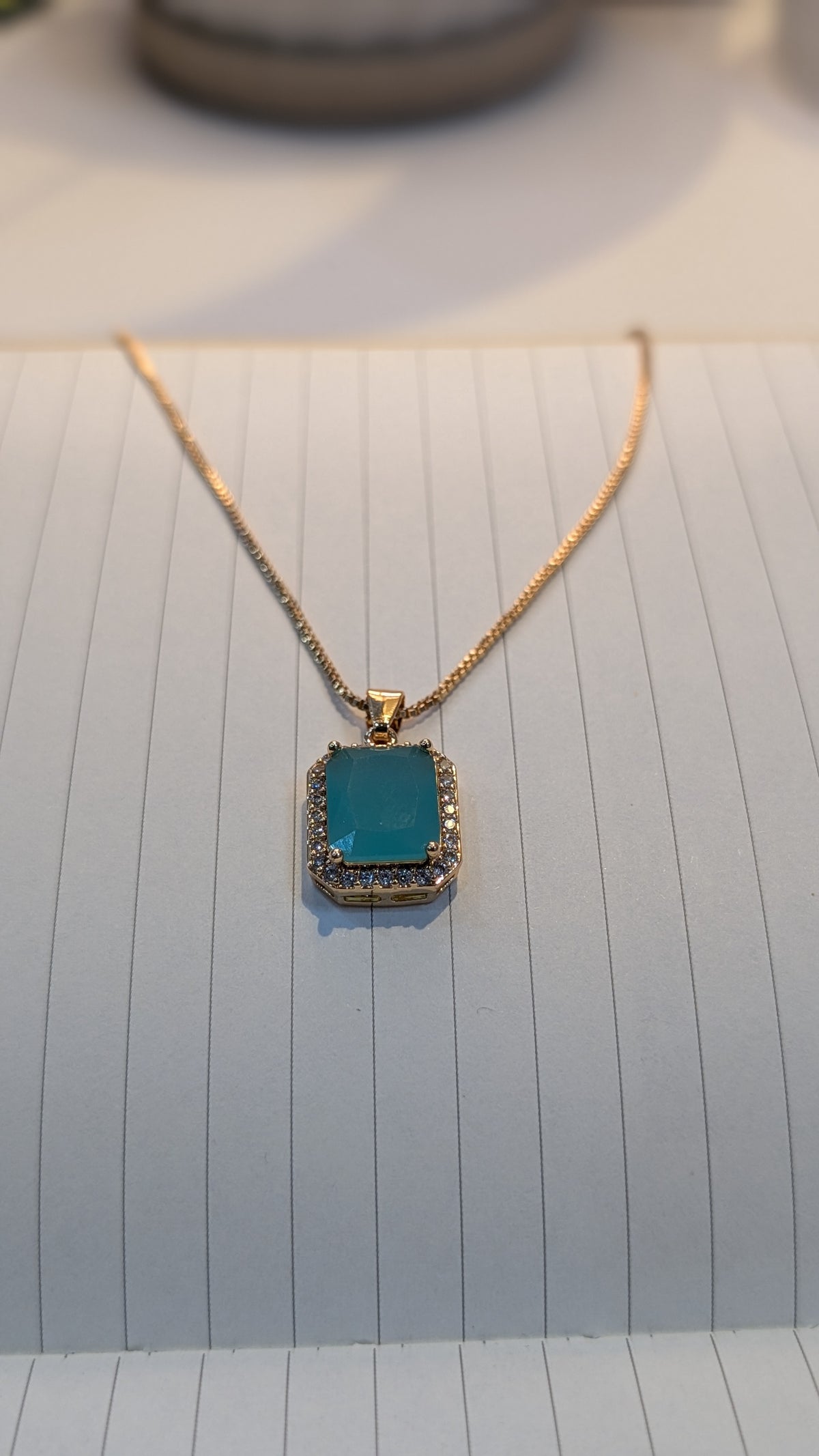 Gold-tone chain necklace with a rectangular teal pendant bordered by small clear stones.