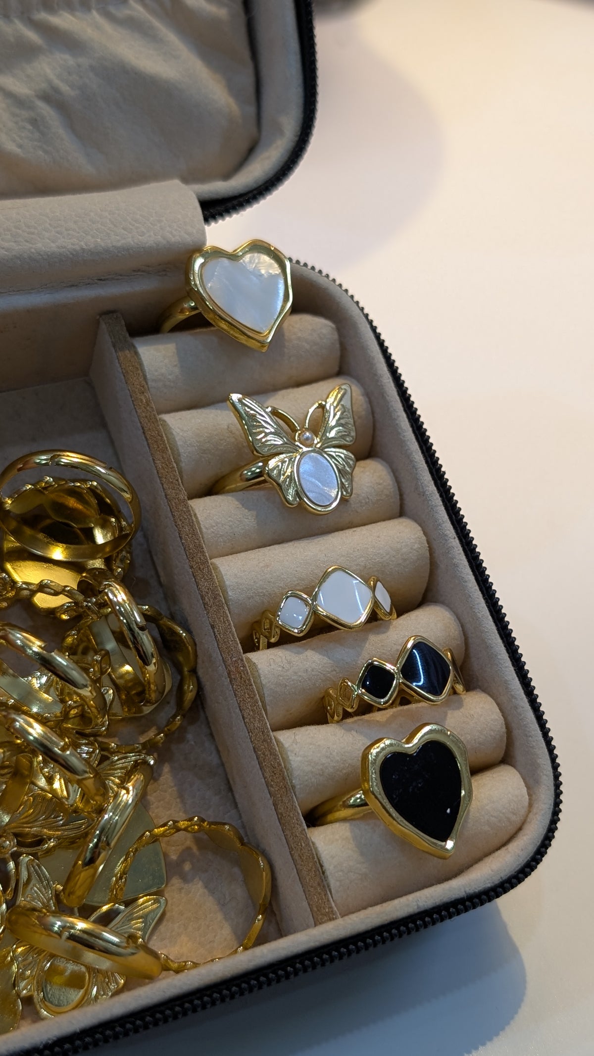 Gold-tone rings in a beige jewelry case, showing heart-shaped light and black faces, a butterfly design with a pale center, and small irregular pale and black insets.