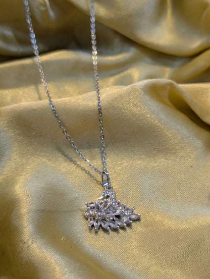 A silver-tone necklace with a fan-shaped cluster pendant made of clear faceted stones, displayed on a draped gold-colored fabric.