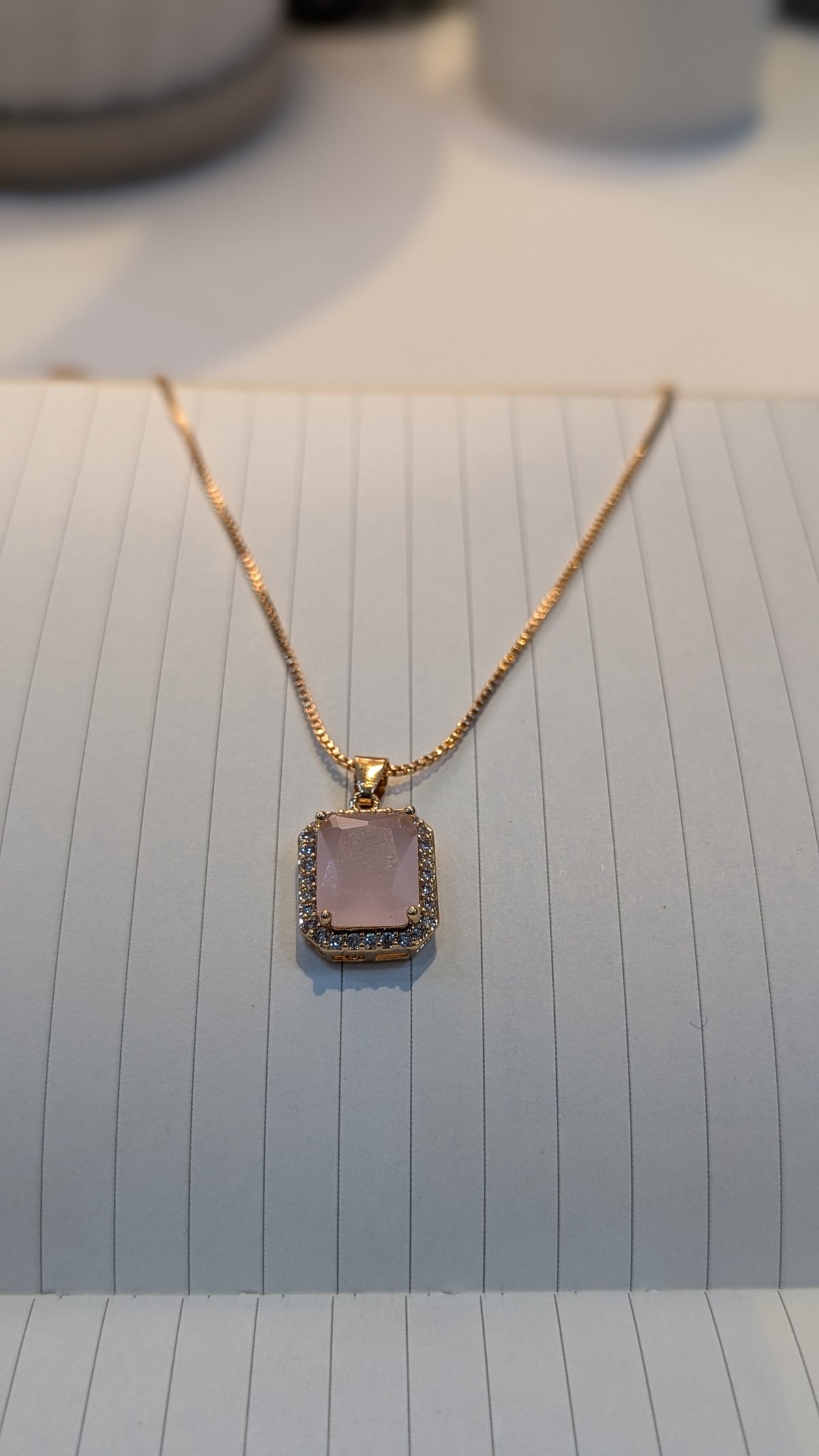 A thin gold-tone chain necklace displaying a rectangular pendant with a pale pink center and a halo of small clear stones.