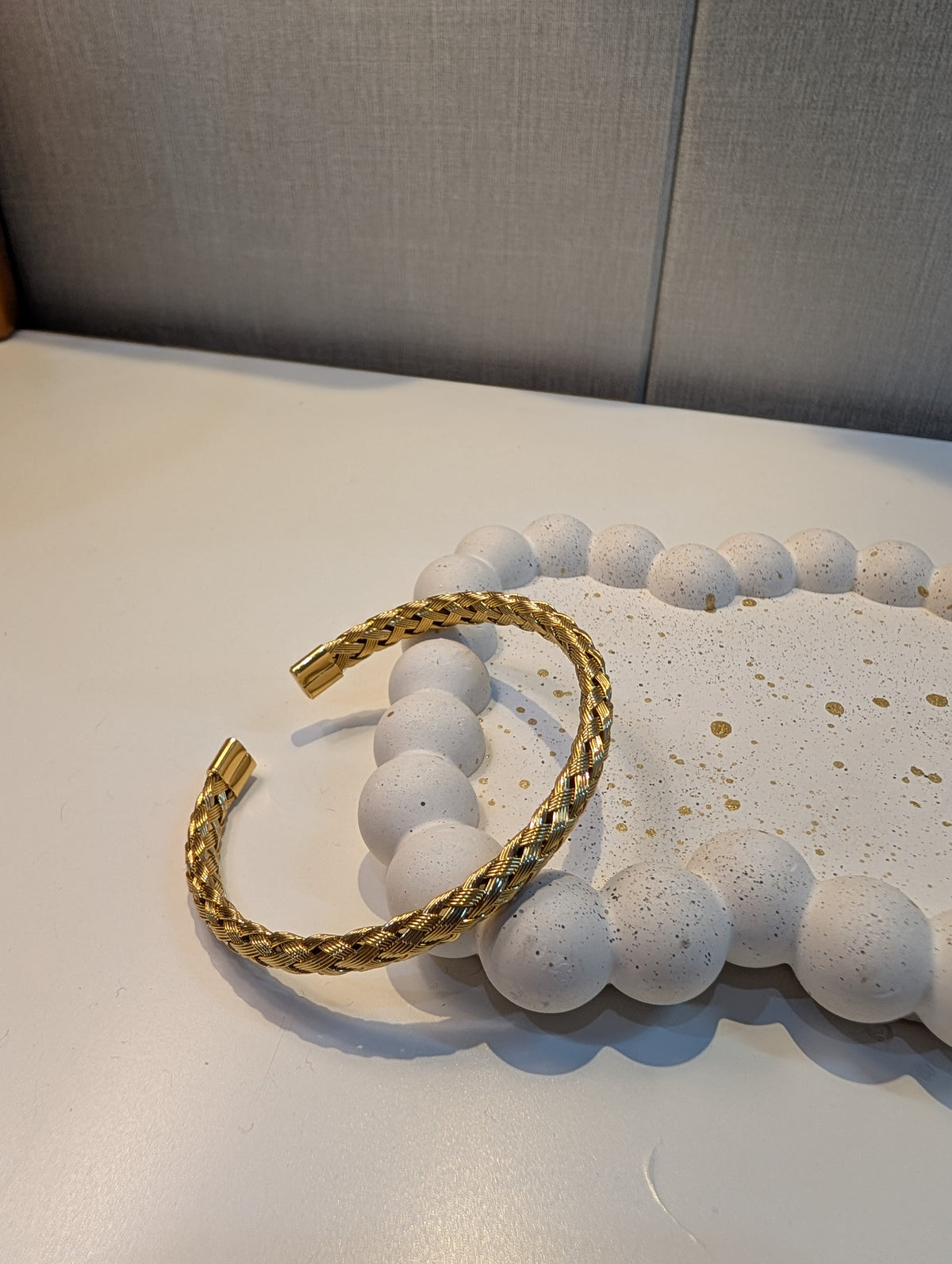 Gold-tone open cuff bracelet with a braided twist texture and smooth capped ends resting on a white speckled tray.