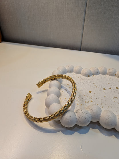 Gold-tone open cuff bracelet with a braided twist texture and smooth capped ends resting on a white speckled tray.