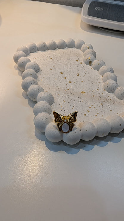 Gold-tone butterfly ring with textured white wings and a pale purple oval center, resting on a white speckled dish.