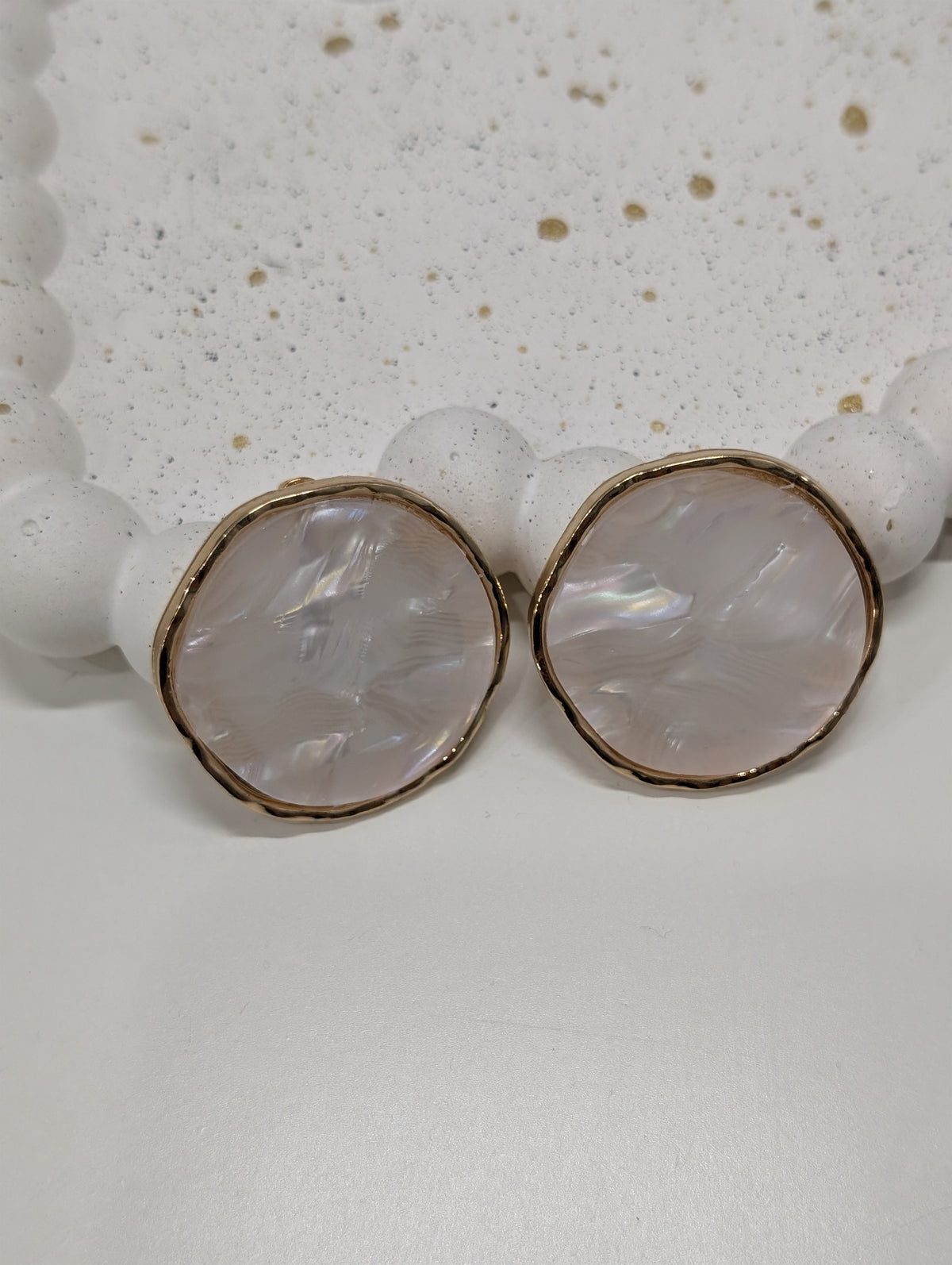 Two round pearlescent pale pink earrings with wavy gold-tone rims, lying side by side on a white surface.