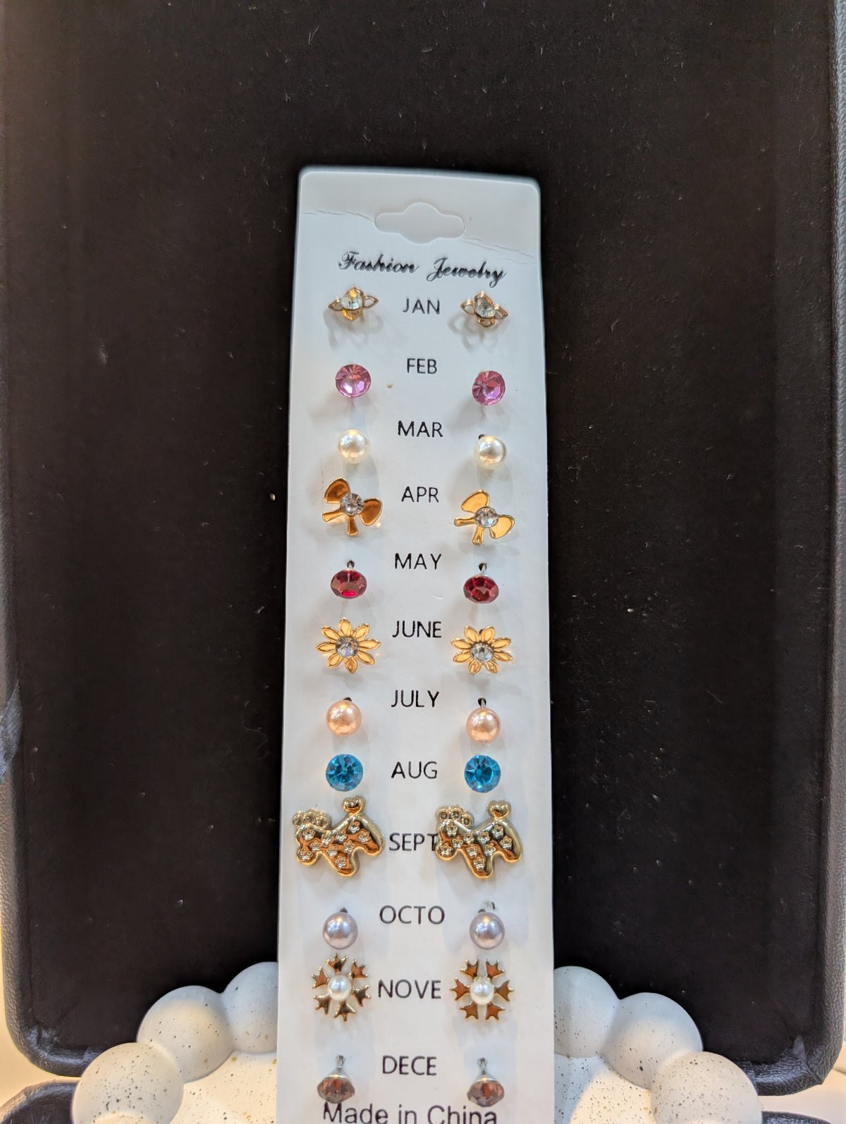A white display card holding a set of small stud earrings arranged vertically by month. The designs feature stars, flowers, bows, and simple stones in assorted colors like pink, clear, red, orange, blue, and purple.