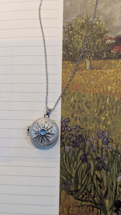 A silver-tone necklace featuring a fine chain and a round locket pendant with an engraved sunburst design and a central blue accent.