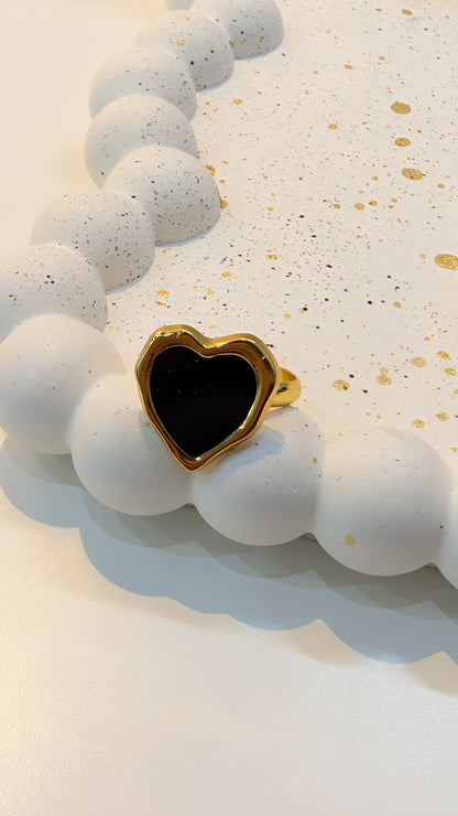 Gold-tone ring with a black heart-shaped center resting on a white speckled tray with rounded bead edging.