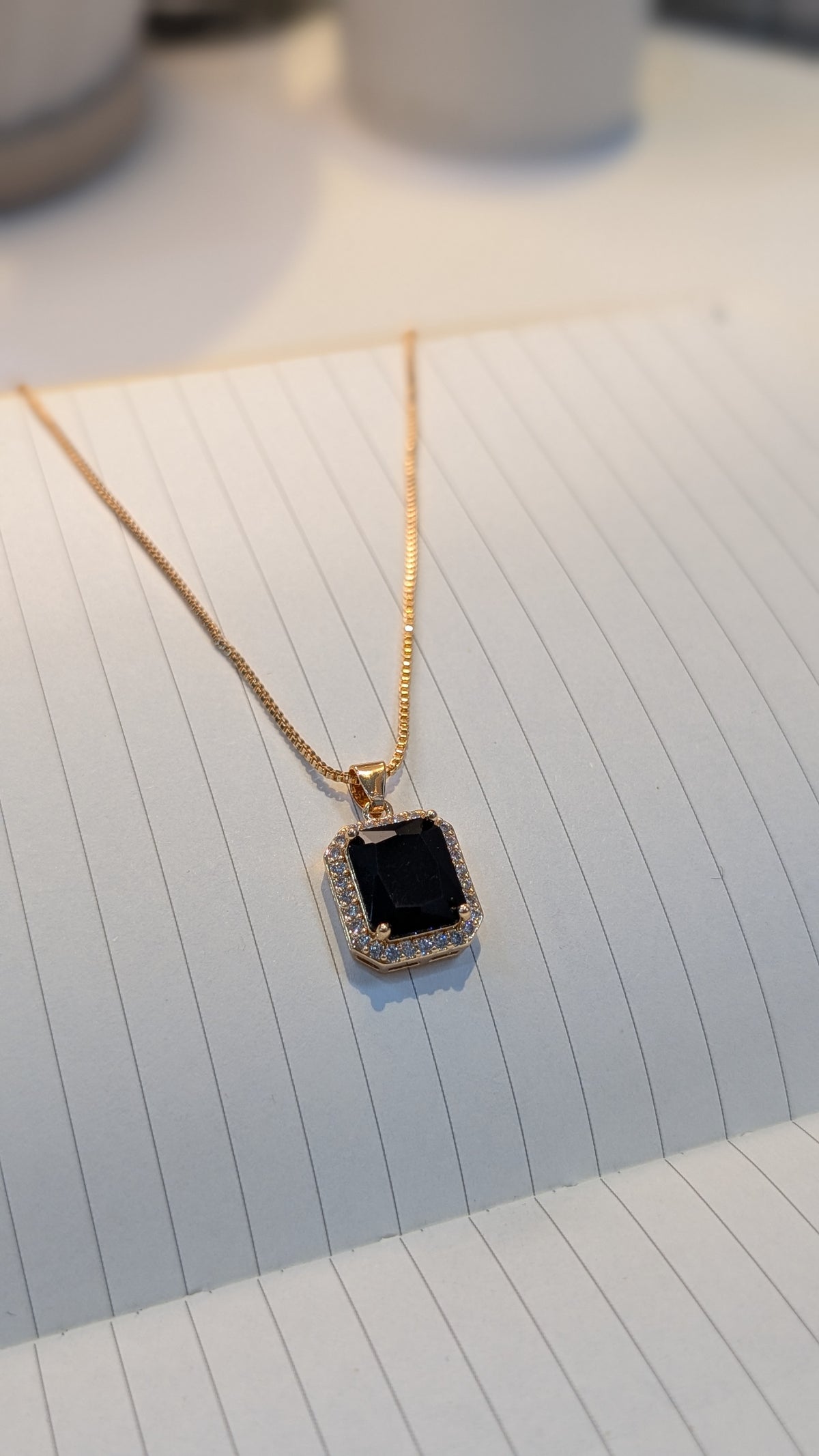 A thin gold-tone chain necklace with a rectangular pendant. The pendant has a large black center surrounded by a halo of small, clear stones, resting on a lined white notepad.