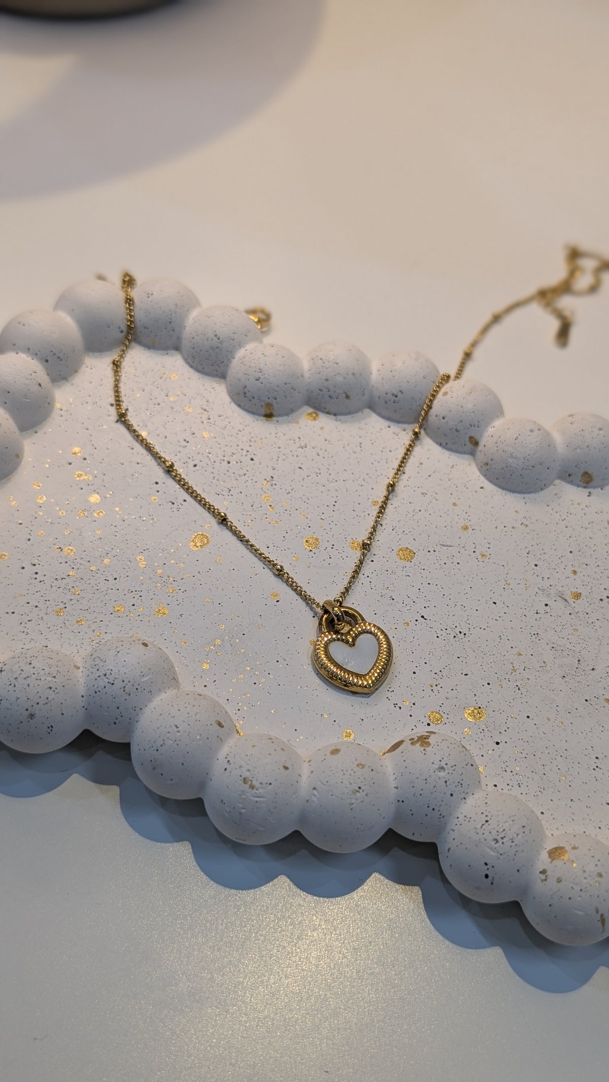 A fine gold-tone chain necklace with an open heart pendant that has a textured border, displayed on a speckled white tray.