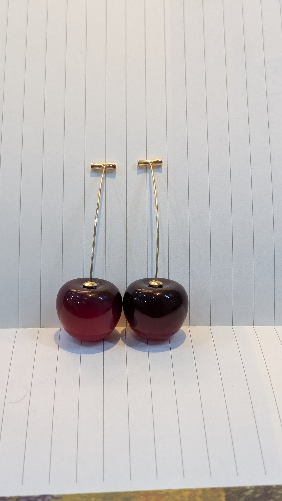A pair of drop earrings with long gold-tone stems and glossy dark red spherical charms, shown standing upright against a lined white background.