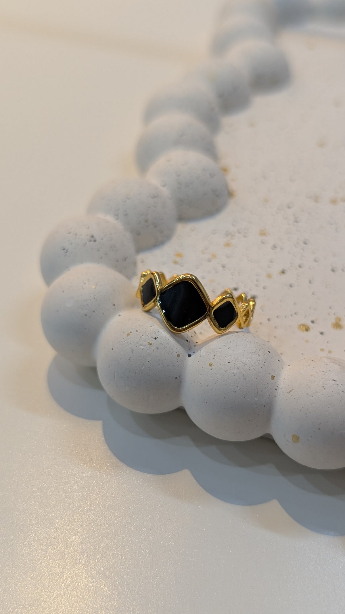 A gold-tone ring with a central black kite-shaped setting and two smaller black square settings on either side, resting on a textured white surface.