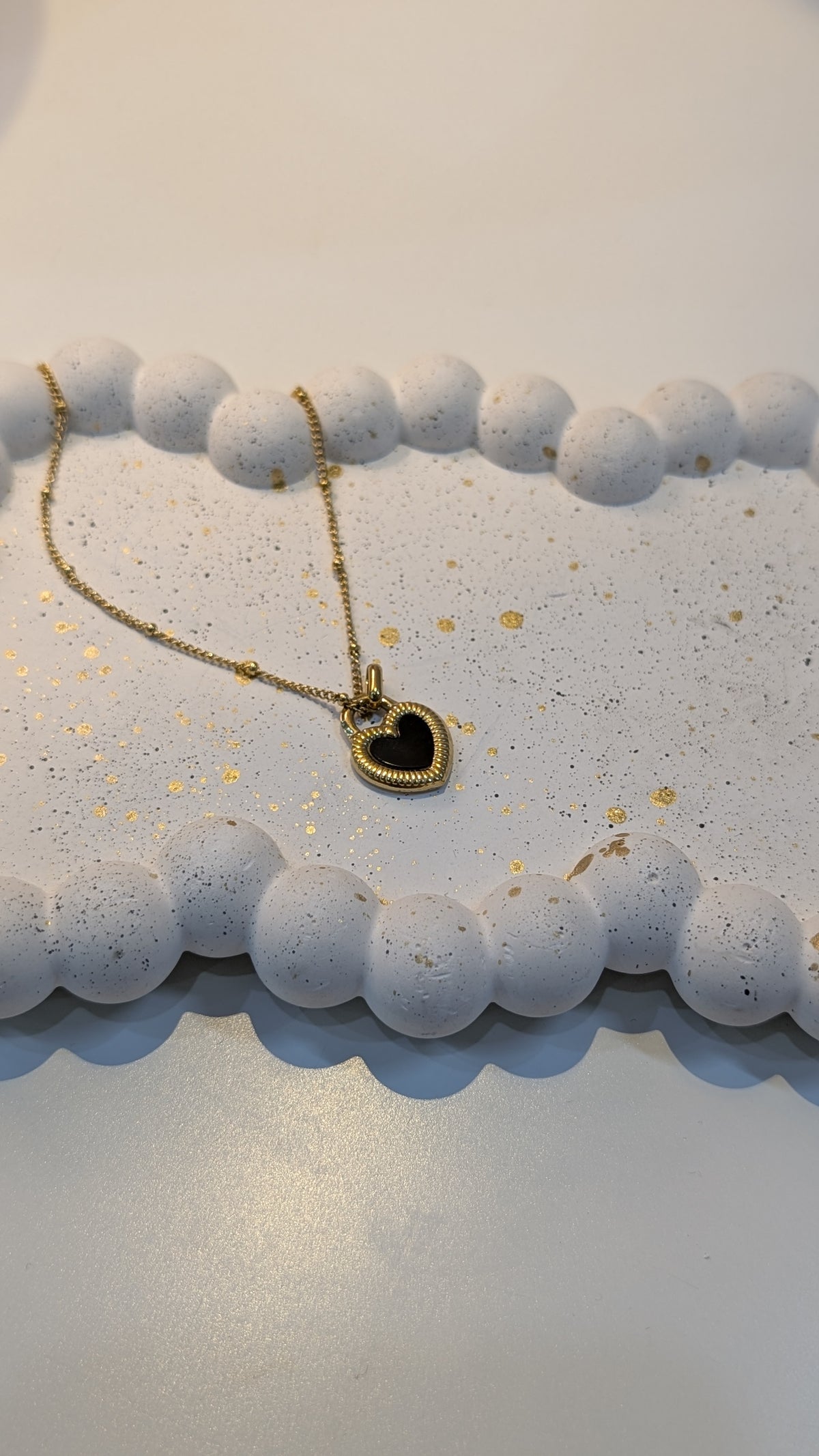 A thin gold-tone chain necklace with a heart pendant that has a black center and a textured gold-tone border, resting on a white speckled tray with a beaded rim.