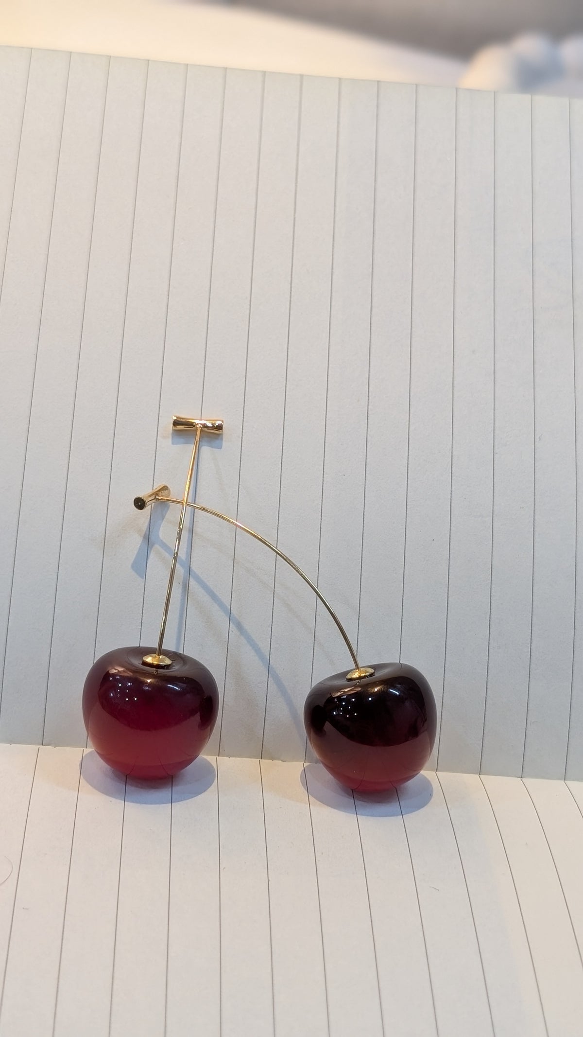 Pair of cherry-shaped earrings with glossy dark red round bodies and gold-tone stems, resting on lined paper.