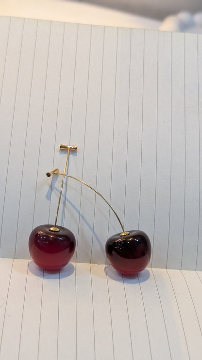 Pair of cherry-shaped earrings with glossy dark red round bodies and gold-tone stems, resting on lined paper.