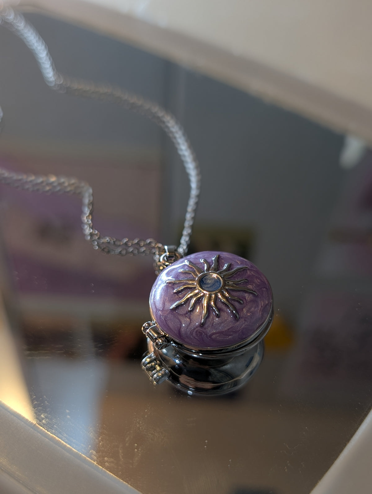 A fine silver-tone chain holds a circular pendant with a domed, textured purple top. A raised gold-tone sunburst motif sits in the center with a small dark blue circular detail. The pendant base appears to be a hinged box style in silver-tone metal.