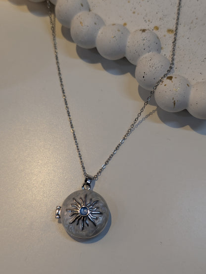 Silver-tone necklace with a fine cable chain and a round pendant decorated with a raised sunburst design around a small blue centre stone, shown on a light surface.