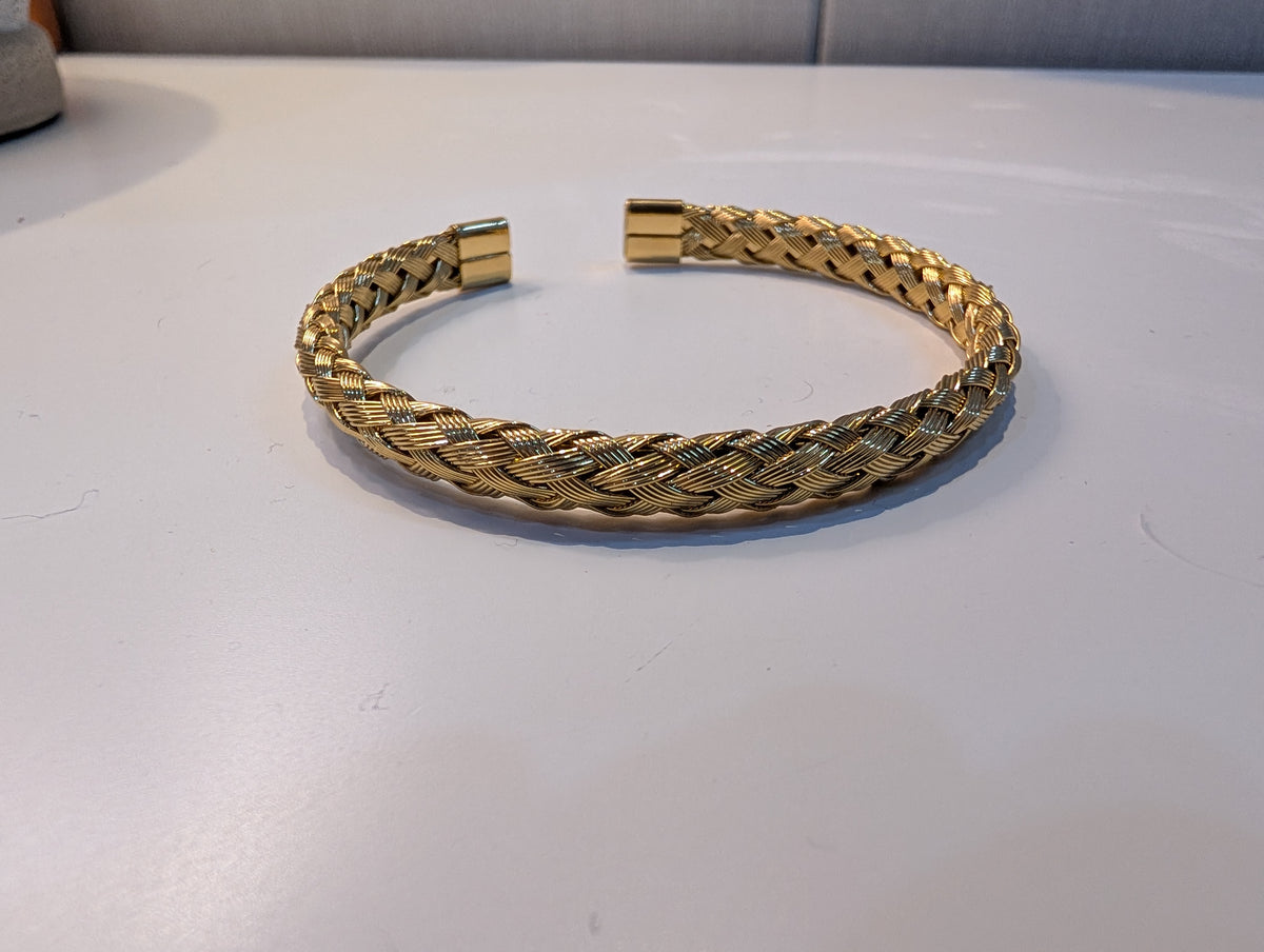 A gold-tone open cuff bracelet with a textured braided design and flat squared ends, lying on a white surface.