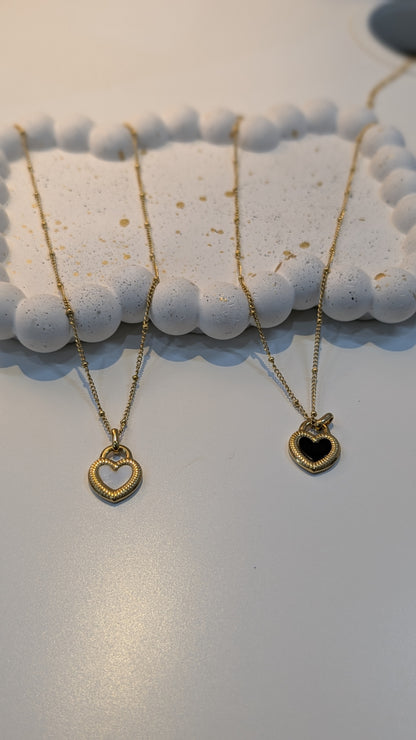 Two gold-tone box chain necklaces with heart pendants, one with a light center and one with a dark center, resting on a textured white tray.