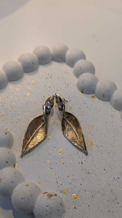 Pair of gold-tone leaf drop earrings with textured vein lines and polished curved tops, photographed on a speckled surface.