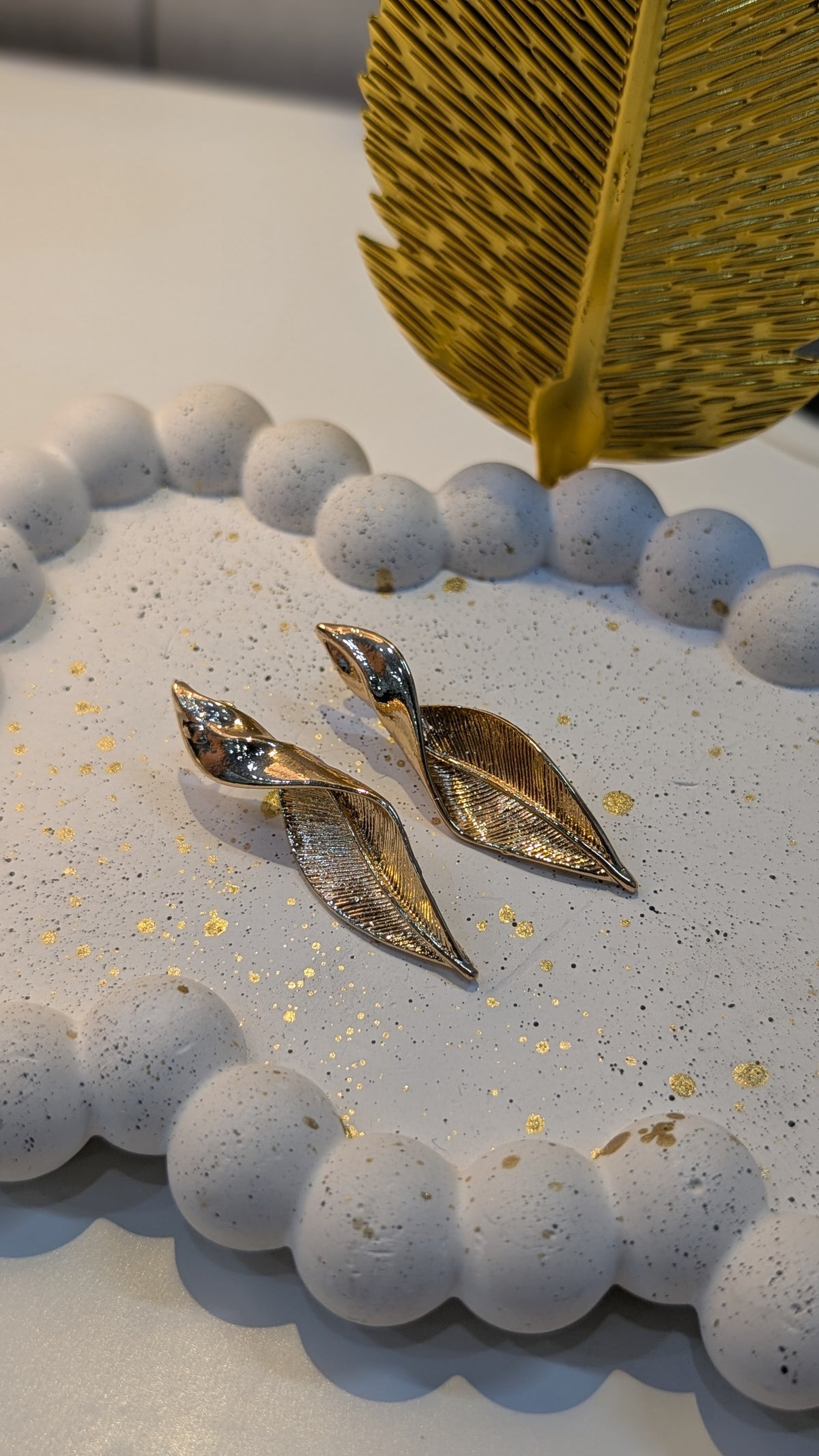 Gold-tone leaf-shaped earrings with textured engraved surfaces displayed on a white speckled tray.