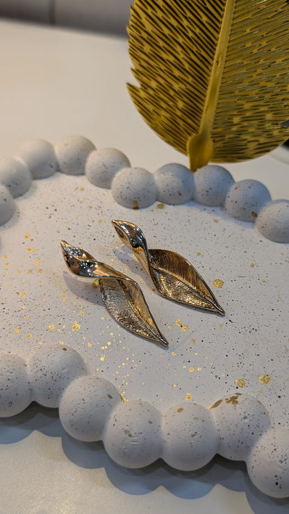 Gold-tone leaf-shaped earrings with textured engraved surfaces displayed on a white speckled tray.