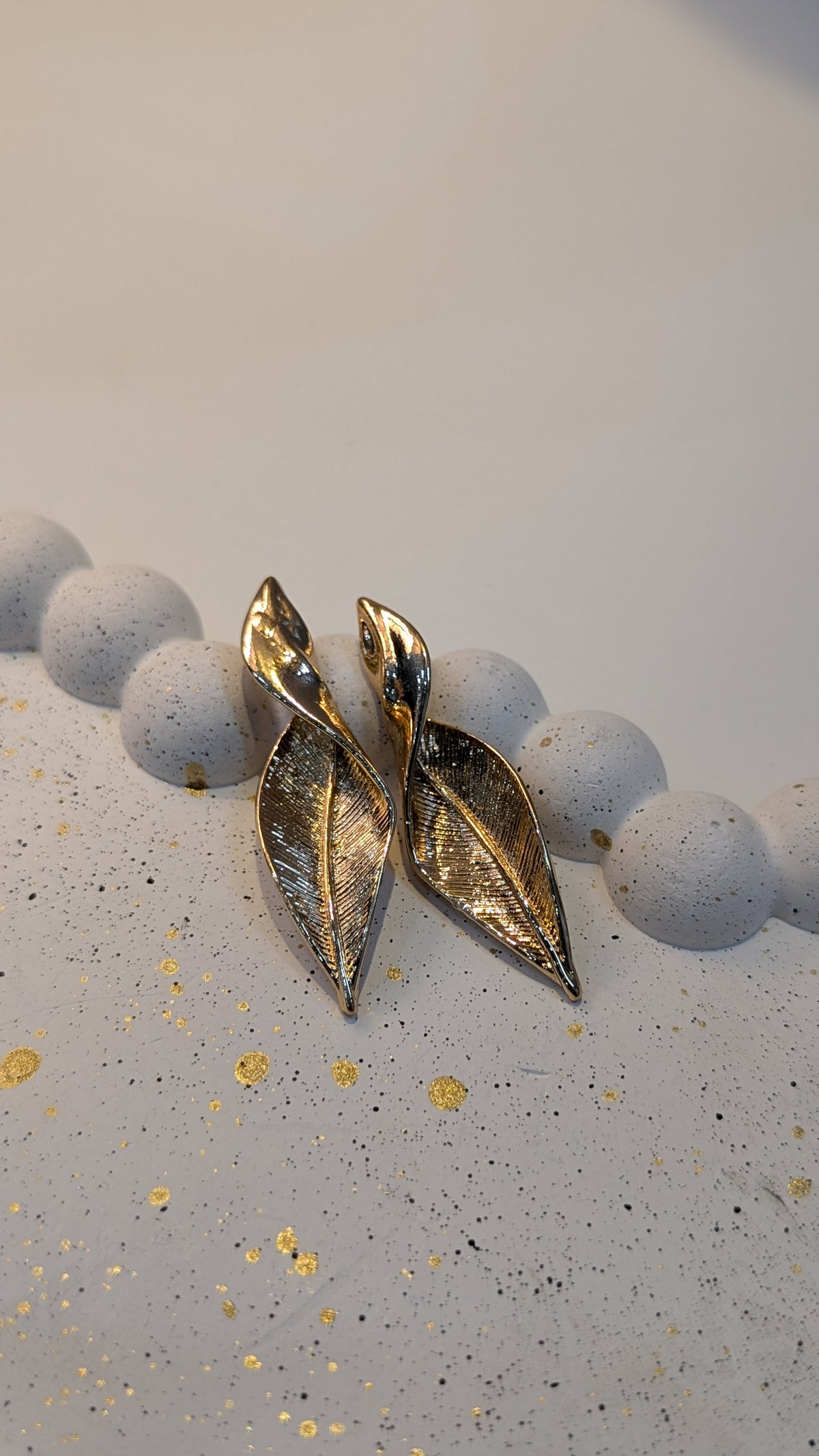 A pair of gold-tone leaf-shaped earrings with engraved vein texture and a polished finish, resting on a pale speckled surface.