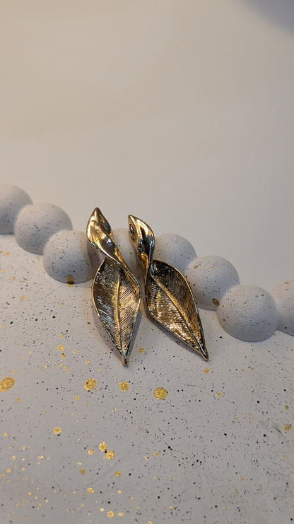 A pair of gold-tone leaf-shaped earrings with engraved vein texture and a polished finish, resting on a pale speckled surface.