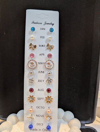 White display card holding month-themed stud earrings labeled JAN through DEC, showing mixed shapes and colors including blue, white, pink, red, orange, and clear accents with gold-tone and silver-tone settings.