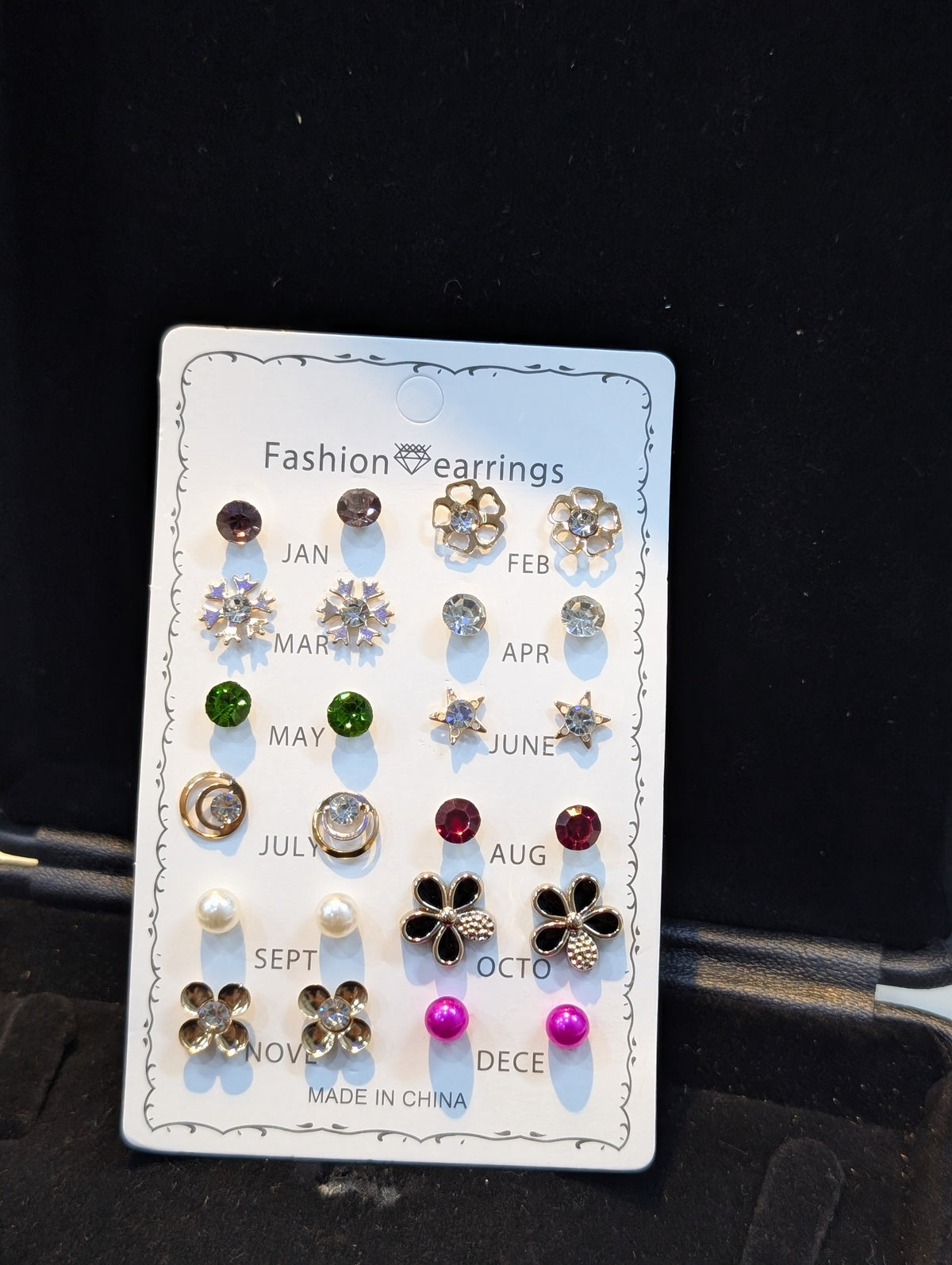 Assorted small stud earrings on a white month-labeled display card, including flower, star, and round designs in various colors with gold-tone and silver-tone settings.