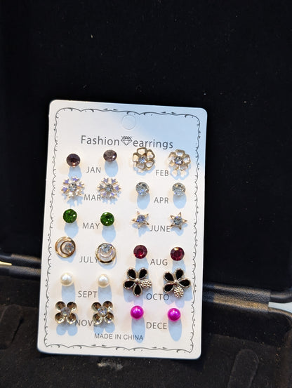 Assorted small stud earrings on a white month-labeled display card, including flower, star, and round designs in various colors with gold-tone and silver-tone settings.