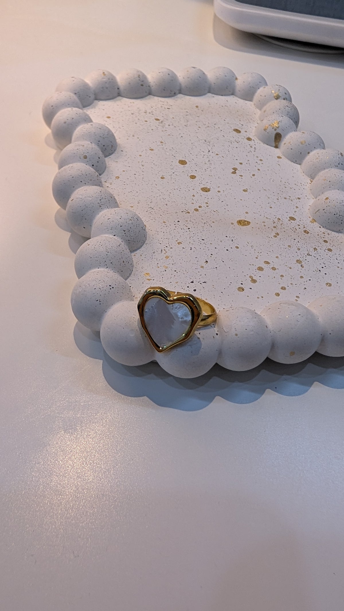 A gold-tone ring with a smooth, pale blue heart-shaped center, resting on a white speckled tray.