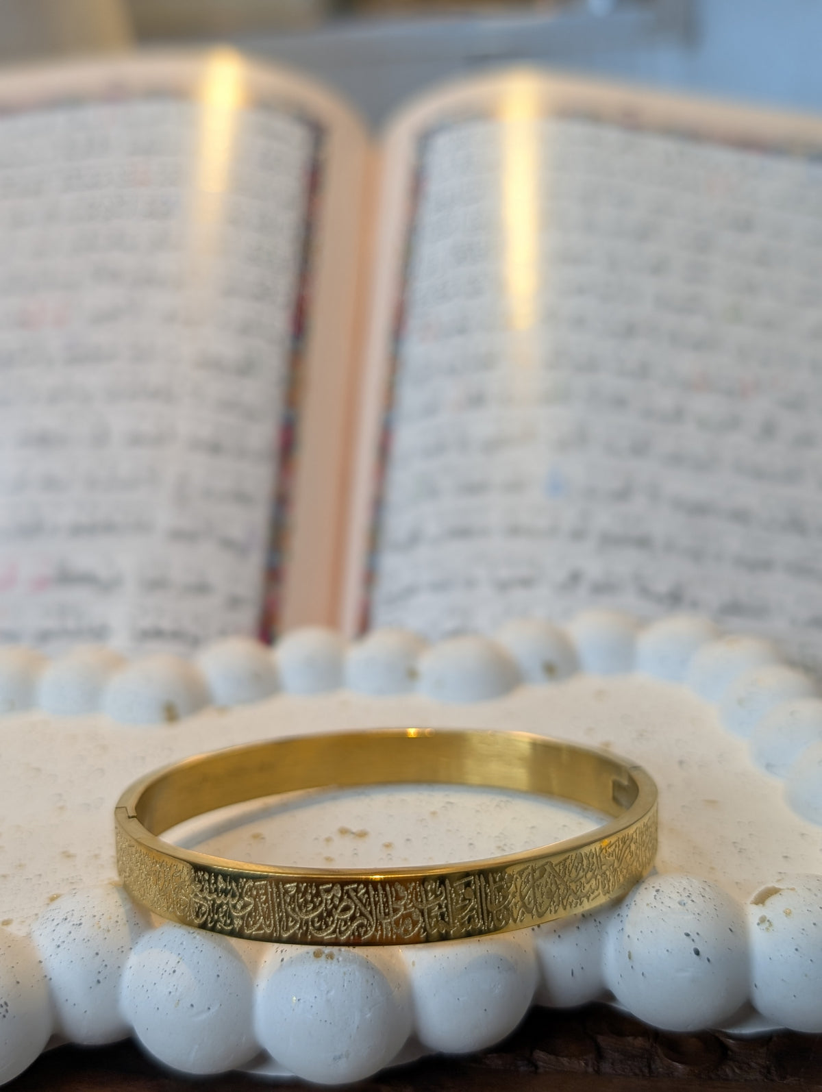 A gold-tone band ring with an intricate engraved pattern, resting on white beads in front of a softly blurred open book.