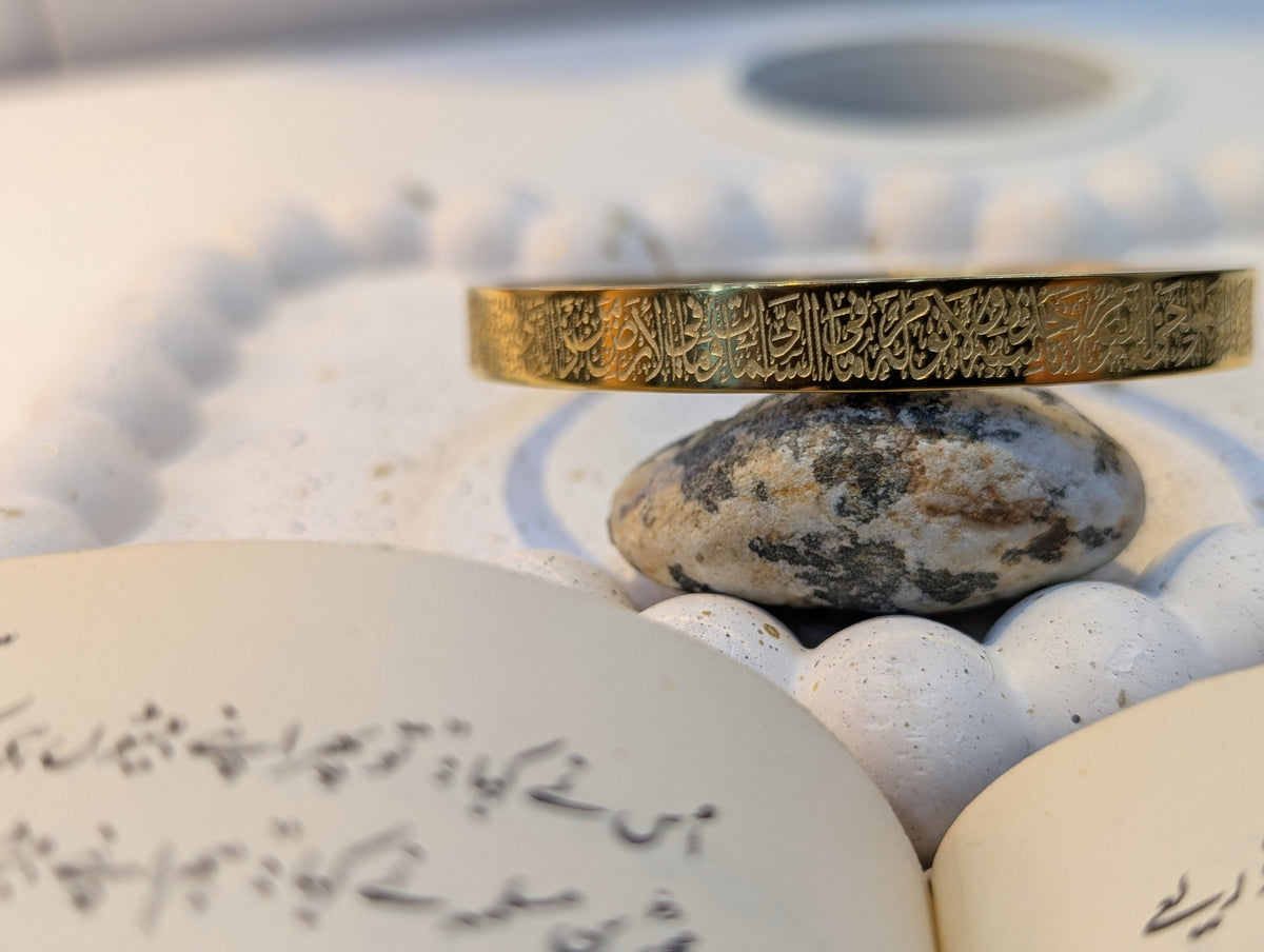 A wide gold-tone band ring with intricate engraved calligraphy script, resting on a speckled stone.